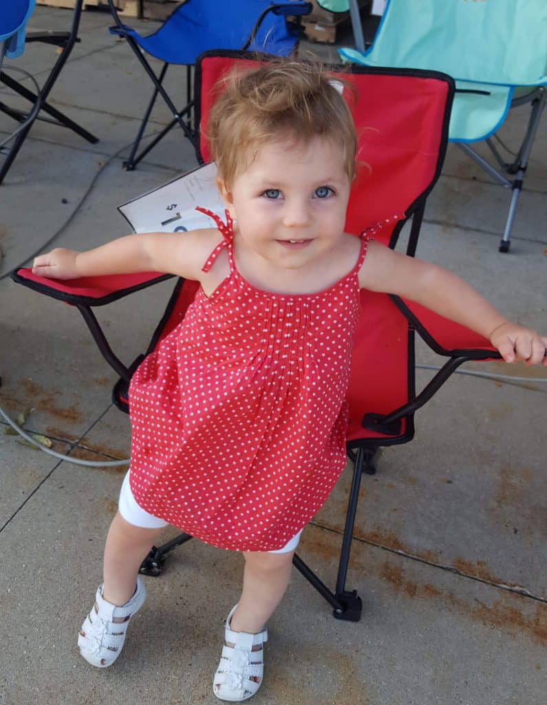 one year old in a red dress standing in front of a red fold-up chair